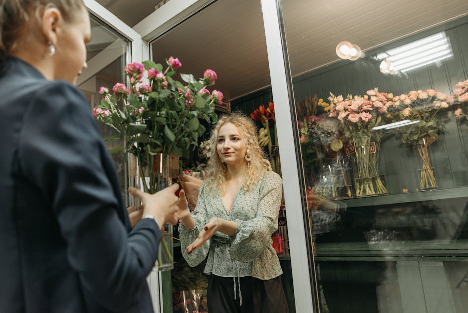 A woman with curly blonde hair, dressed in a patterned blouse, is receiving a bouquet of pink roses from a florist dressed in a dark jacket inside a floral shop. The bouquet is arranged with lush gree
