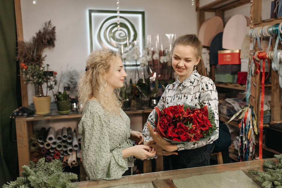 A young woman with light brown hair tied back, wearing a floral patterned shirt, is receiving a bouquet of fresh red roses, red lilies, and green foliage from a florist with curly blonde hair dressed