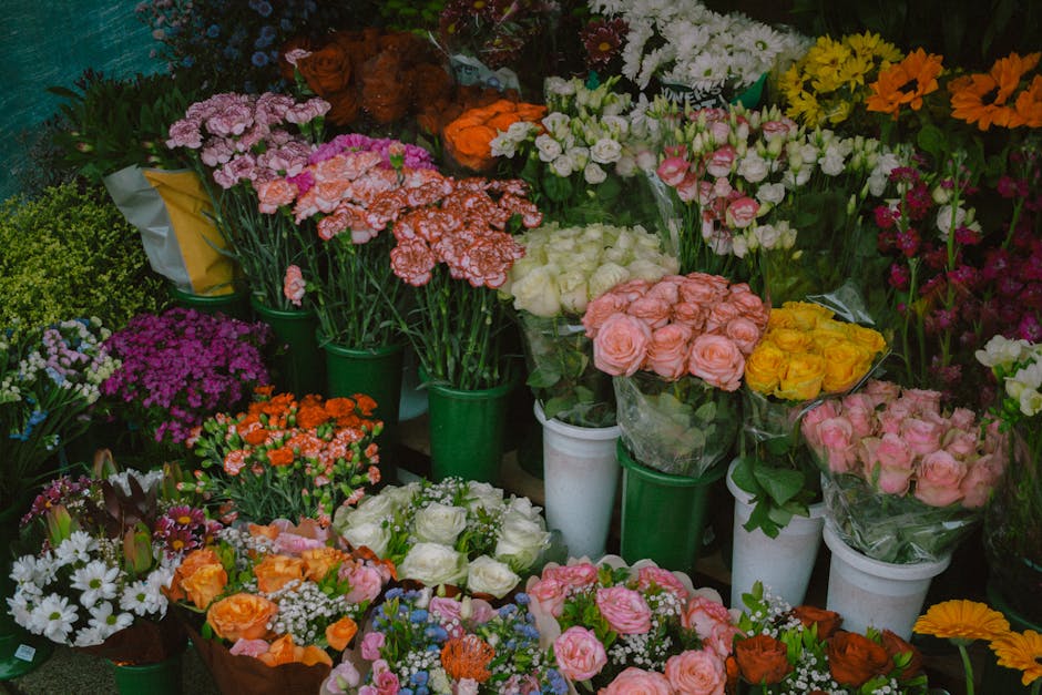 A vibrant display of fresh flowers arranged in various bouquets and containers, including roses in shades of pink, yellow, and white, alongside clusters of purple, orange, and white blossoms. The flow