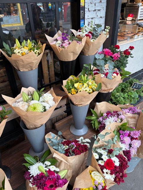 A display of fresh floral bouquets arranged in tall, tapered grey vases and wrapped in brown paper, featuring a variety of flowers including white lilies, yellow daisies, purple and pink carnations, a