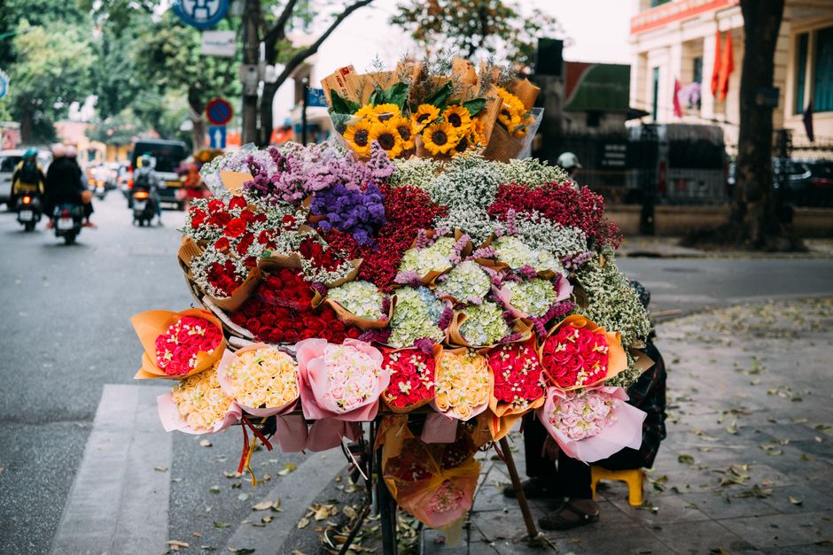 A vibrant flower stall situated on a city street, featuring a large, multi-layered bouquet of fresh flowers arranged on a bicycle cart. The arrangement includes bright yellow sunflowers, clusters of p
