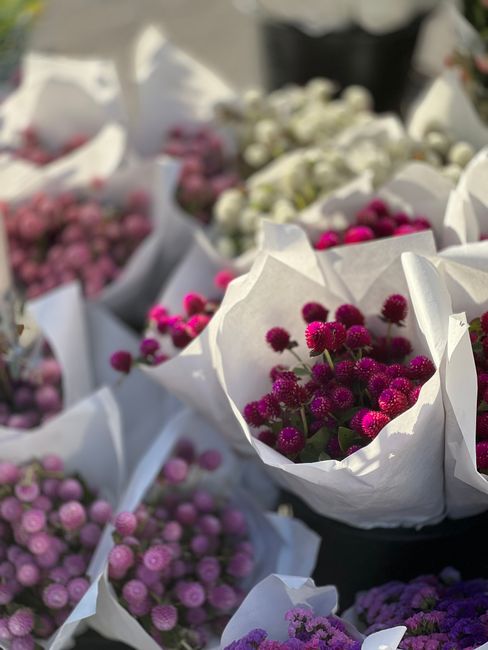 A display of fresh flowers arranged in white paper wrapping, featuring vibrant magenta and purple blooms with round, clustered petals, situated outdoors on a bright day. The flowers are arranged in mu