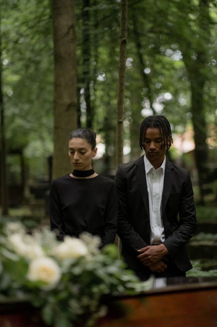 A somber scene featuring two individuals in formal black attire standing outdoors among tall, leafy trees. The woman on the left wears a black dress with long sleeves and a choker, her hair pulled bac