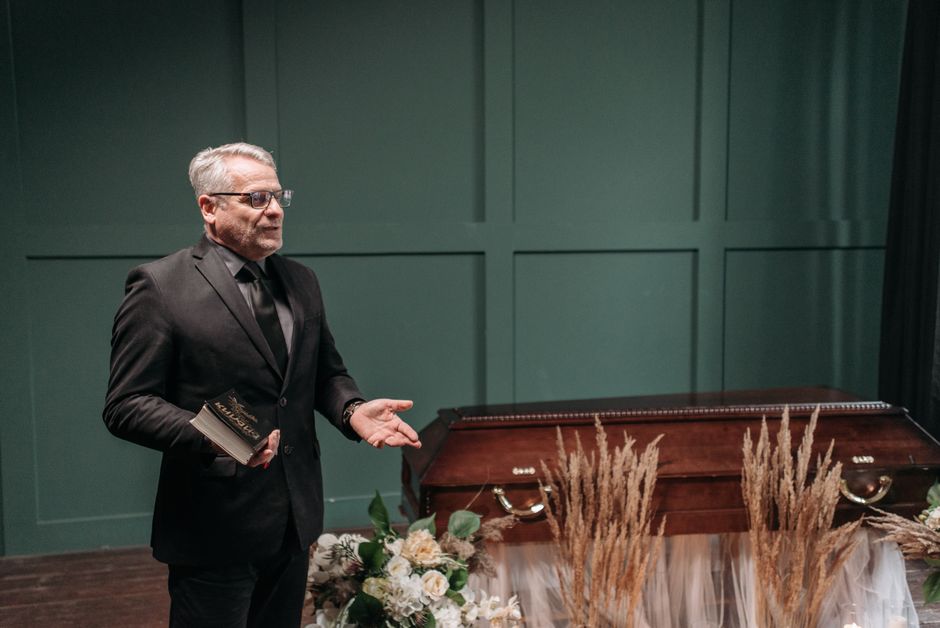 A man in a dark suit and glasses stands beside a wooden casket adorned with floral arrangements at a funeral service. The floral display includes white roses, cream-colored peonies, and greenery, arra